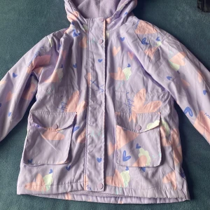 Lila mönstrad jacka med huva - Säljer en söt lila jacka med huva och ett färgglatt mönster av hjärtan och abstrakta former i rosa, blått och grått. Jackan har långa ärmar och praktiska fickor framtill. Perfekt för en färgglad stil!