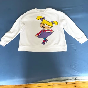 Vit sweatshirt med tecknad figur - Säljer en vit sweatshirt med en stor färgglad tecknad figur på framsidan. Tröjan har långa ärmar och en rund halsringning. Perfekt för en avslappnad stil med en rolig twist!