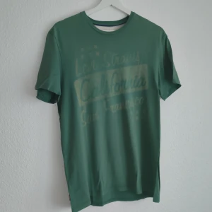 Grön t-shirt från Levi Strauss & Co - Säljer en grön t-shirt från Levi Strauss & Co med tryck på framsidan. T-shirten har en rund hals och korta ärmar. Perfekt för en avslappnad stil.