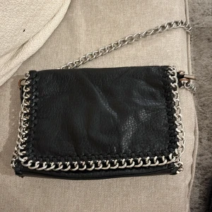 Svart axelväska med kedjedetaljer - Snygg svart axelväska från Liamo med silverfärgade kedjedetaljer runt kanterna. Väskan har en dragkedja upptill och en extra ficka med dragkedja på baksidan. Perfekt för att ge en edgy touch till din outfit. Den är sönder på baksidan därav priset men det syns mer på kamera än i verkligheten! 