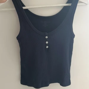 Svart ribbad linnetopp från Brandy Melville - Snygg svart ribbad linnetopp från Brandy Melville med knappar framtill. Perfekt för en stilren och bekväm look. Passar till många olika outfits och tillfällen.