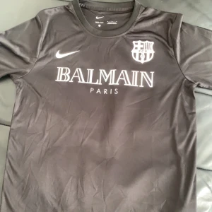 Balmain barcelona fotbollströja - Snygg svart fotbollströja från Nike med Balmain Paris-tryck och FCB-logga på bröstet. Tillverkad i Dri-FIT-material för optimal komfort. Perfekt för både träning och vardag.