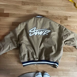 Beige bomberjacka med broderad text  - Säljer en beige bomberjacka från Stwnd med broderad text på ryggen och årtalet 1997 på framsidan. Jackan har långa ärmar och ribbade muddar i svart och vitt. Perfekt för en cool och avslappnad stil.