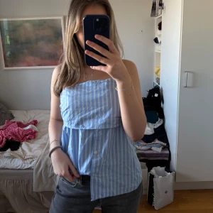 Blå- och vitrandig tubtopp - Säljer en söt blå- och vitrandig tubtopp med knappar framtill. Perfekt för sommardagar och ger en fräsch look. Toppen är ärmlös och har en avslappnad passform. Den har ett superlitet hål som inte syns