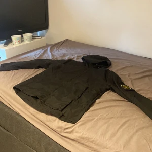 Svart jacka med huva stone islnad - Säljer en svart jacka med huva och dragkedja. Jackan har en logga på ärmen och är perfekt för både höst och vår. Den har en stilren design och är lätt att matcha med olika outfits.