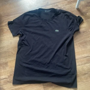 Svart t-shirt från Lacoste - Säljer en klassisk svart t-shirt från Lacoste med den ikoniska krokodilloggan på bröstet. T-shirten har en rund halsringning och korta ärmar. Perfekt för en stilren och avslappnad look.