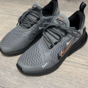 Grå Nike Air Max 270 - Snygga grå Nike Air Max 270 sneakers med orange och vita detaljer. Skorna har en luftig design med snörning och en bekväm sula för extra komfort. Perfekta för både träning och vardag.