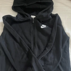 Svart hoodie från Nike - Säljer en svart hoodie från Nike med dragkedja och broderad logga på bröstet. Hoodien har långa ärmar och en klassisk huva. Det är Large barn så typ 158-164/xs