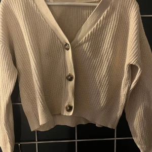 Beige stickad kofta med knappar - Säljer en beige stickad kofta med stora knappar framtill. Koftan har en croppad passform och långa ärmar. Perfekt för en stilren och bekväm look. Passar bra till både jeans och kjol.