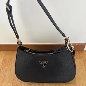 Svart axelväska från Guess - Snygg svart axelväska från Guess med guldfärgade detaljer och en praktisk dragkedja. Väskan har en elegant design med Guess-logotypen på framsidan. Perfekt för att bära dina nödvändigheter med stil. Orginalpris 1090 kr 