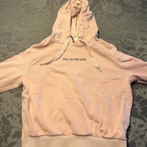 Rosa hoodie med text - Säljer en mysig rosa hoodie med texten 'YOU GLOW GIRL' på bröstet. Den har en justerbar huva med snören och ribbade muddar vid ärmslut och nederkant. Perfekt för en avslappnad stil.