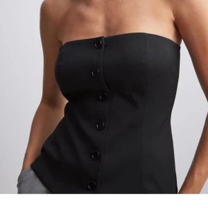 Svart axelbandslös korsettopp - Snygg svart axelbandslös topp med knappar framtill. Perfekt för en stilren look, passar till allt. Toppen har en figurnära passform som framhäver silhuetten.  Slutsåld i alla storlekar med hög efterfrågan! Säljer endast då jag råkade köpa 2! 