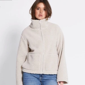 Beige teddyjacka med dragkedja - Mysig beige teddyjacka med hög krage och dragkedja framtill. Perfekt för kyliga dagar med sin fluffiga och varma design. Jackan har en avslappnad passform och långa ärmar.