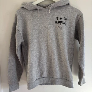 Grå hoodie - Säljer en grå hoodie med texten 'JE M'EN AMOUR' och en stjärna tryckt på både fram- och baksidan. Tröjan har en klassisk huva med dragsko och långa ärmar med ribbade muddar. Perfekt för en avslappnad stil.🩷
