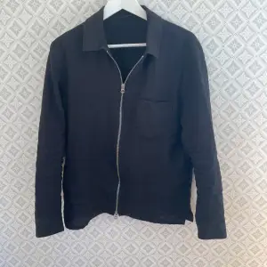 Säljer en stilren overshirt  från J.Lindeberg med dragkedja framtill och en bröstficka. Jackan har en klassisk krage och långa ärmar, perfekt för en minimalistisk look.