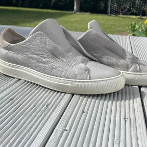 Grå sneakers från Axel Arigato - Snygga grå sneakers från Axel Arigato i skinn. De har en stilren design utan snörning och en rund tå. Perfekta för en avslappnad stil.