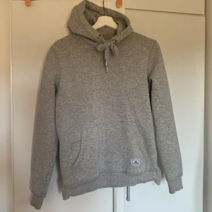 Grå hoodie från Tenson - Säljer en stilren grå hoodie från Tenson med justerbar huva och känguruficka. Tröjan har ett stort Tenson-tryck på ryggen och är perfekt för en avslappnad look. Passar bra till både jeans och träningsbyxor.