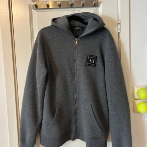 Grå hoodie från Armani Exchange - Säljer en stilren grå hoodie från Armani Exchange med dragkedja och en liten logotyp på bröstet. Perfekt för en avslappnad look. Den har en bekväm passform och är tillverkad i mjukt material.