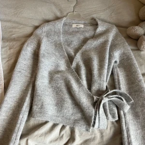 Grå omlottkofta från Gina  - Säljer en mysig grå omlottkofta från Gina. Koftan har långa ärmar och ett knytband i midjan för en justerbar passform. Perfekt för kyliga dagar och att mysa i hemma. 🩶