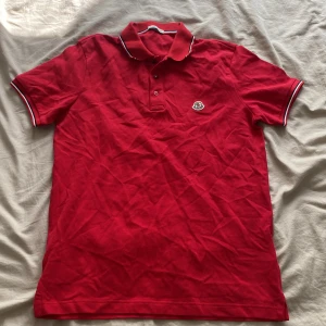 Röd pikétröja från Moncler - Snygg röd pikétröja från Moncler med klassisk krage och knäppning framtill. Tröjan har Moncler-logga på bröstet och vita detaljer på krage och ärmslut. Tillverkad i mjuk bomull, perfekt för en stilren och sportig look.