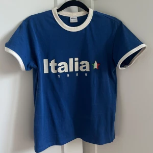 Blå Italia t-shirt  - Blå t-shirt från Bershka. Har några brun-utan-sol-fläckar på kragen på insidan men är rätt säker på att det försvinner när jag tvättar den. Inget som syns när man har på sig den. Använd max 5 gånger. Ord. Pris: 139 kr.