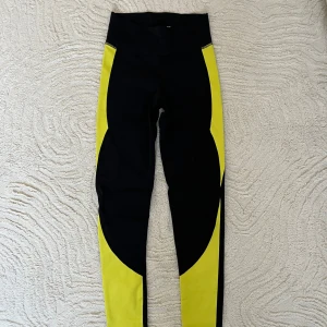Svarta och gula träningsbyxor från NA - Säljer ett par träningsbyxor i svart och gult från NA. De är supersköna med coola detaljer på sidorna av benen och gjorda i ett stretchigt material som passar perfekt för träning. De har även reflekterande detaljer längst ner baktill på benen vilket gör de synliga.  