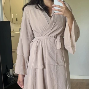 Beige omlottklänning med bälte - Säljer en beige omlottklänning med vida ärmar och knytband i midjan. Klänningen har en avslappnad passform och är perfekt för dig som gillar en stilren och enkel look. Materialet känns lätt och luftigt, vilket gör den skön att bära.