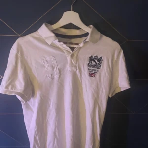 Vit pikétröja från Morris XS - Vit pikétröja från Morris i klassisk modell med broderat emblem och brittisk flagga på bröstet. Tröjan har krage, korta ärmar och en broderad siffra 9 på ena sidan. Perfekt för en preppy stil och tillverkad i bomull.