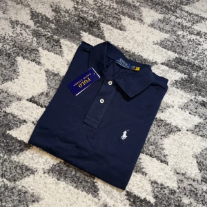 Ralph Lauren Pike - Klassisk navy pikétröja från Ralph Lauren med vit broderad logga på bröstet. Tröjan har korta ärmar, krage och två knappar framtill. Tillverkad i bomull för en skön och luftig känsla. Perfekt för en stilren och avslappnad look.