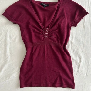 Vinröd topp - Supersnygg vinröd topp från Morgan med djup v-ringning och coola silvriga nitar framtill. Modellen är figurnära med ribbade muddar vid ärmslut och midja. Kortärmad och stretchig. I princip aldrig använd.