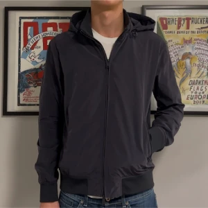 Svart vindjacka från Armani  - Snygg svart vindjacka med minimalistisk design och hel dragkedja framtill. Jackan har långa ärmar, ribbade muddar och två sidofickor med dragkedja. Perfekt för dig som gillar en clean och stilren look.