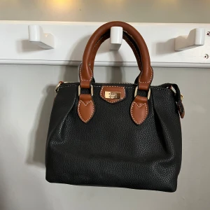 Liten svart och brun handväska - Snygg svart handväska från Feel i skinn med bruna handtag och detaljer. Väskan har guldfärgad dragkedja och ett blommigt innerfoder. Perfekt liten väska för det viktigaste. 