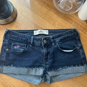 Mörkblå jeansshorts från Hollister - Snygga mörkblå jeansshorts från Hollister med råa kanter och uppvikta benslut. Klassisk femficksmodell med broderad logga på fickan och detaljerade bakfickor. Perfekta för varma dagar och en avslappnad stil.