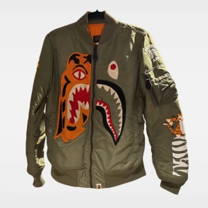 BAPE bomberjacka med tigerpatch - Säljer en grön bomberjacka från BAPE - A Bathing Ape  med ikoniska tiger- och hajpatches framtill. Jackan har orange foder, dragkedja, ärmlogga och coola detaljer på ärmen. Tyvärr hål på insidan där hängflärpen brukar vara. Lite tvättludd på muddar.
