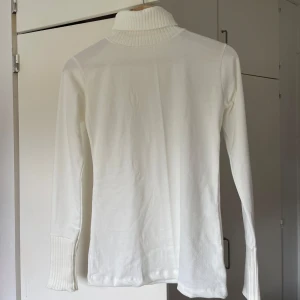 Vit tröja med ribbade detaljer - Klassisk vit tröja med ribbad hög krage och breda ribbade muddar vid ärmsluten. Tröjan är långärmad och har en clean, enkel look som funkar till allt.  perfekt för lager på lager under kyliga dagar.