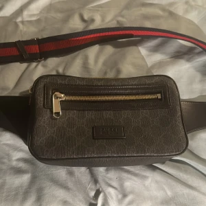 Gucci svart magväska  - Snygg svart magvsäka  från Gucci med klassisk röd och marinblå randig rem. Väskan har guldfärgad dragkedja framtill, präglad logotyp och är gjord i skinn med textilinslag. Perfekt för att bära det viktigaste och ge outfiten en lyxig touch. (Äkta väska)