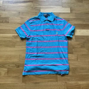 Ralph Lauren piké  - Snygg pikétröja från Ralph Lauren i blått med rosa ränder. Klassisk krage, två knappar och broderad gul logga på bröstet. Tröjan är i bomull och har korta ärmar, perfekt för en chill och stilren look. Har vita fläckar på magen Jag skickar ej bild när jag bär plaggen och jag vet ej vilken längd och vikt de passar då jag inte prövar kläderna jag säljer 😃
