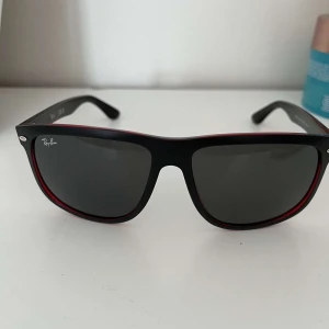 Svarta Ray-Ban solglasögon med röd kant - Säljer mammas Ray-Ban solglasögon i klassisk modell med svarta bågar,  röd kant runt linserna. Mörka glas och plastmaterial. Kommer med originalfodral, putsduk och Ray-Ban certifikat. Som nya, knappt använda då hon inte gillar dom.