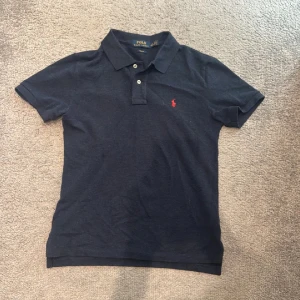 Mörkblå pikétröja från Polo Ralph Lauren - Klassisk mörkblå pikétröja från Polo Ralph Lauren med korta ärmar och röd broderad logga på bröstet. Tröjan har krage, knappar framtill och är tillverkad i mjuk bomull. Perfekt för en stilren och avslappnad look.