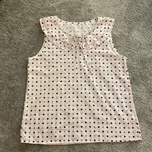 Prickig blus med rosett - Supersöt vit blus med rosa och lila prickar.  Volangkrage. Framtill finns en söt knytrosett vid halsen. Perfekt för dig som gillar en lekfull och trendig stil. Luftigt material som passar bra till varma dagar. 