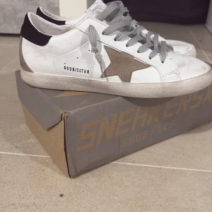 Golden goose superstar skor. - Golden Goose sneakers i vitt skinn med grå mockadetaljer och grå snörning. Sidan pryds av en ikonisk stjärna i beige mocka och bakkappan är svart. Snygg, avslappnad look med platt sula och rund tå. Använde en gång