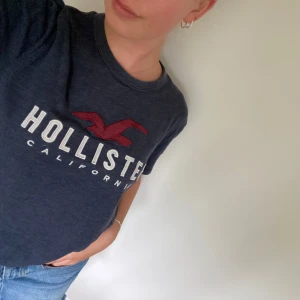 Mörkblå Hollister t-shirt med tryck - Säljer en mörkblå t-shirt från Hollister med klassiskt tryck och röd logga framtill. T-shirten har rund hals, korta ärmar och är gjord i mjuk bomull. Perfekt att styla med en jeanskjol nu till sommaren. 
