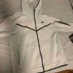 Nike Tech Fleece grå hoodiejacka - Nike Tech Fleece hoodiejacka i ljusgrått med svarta detaljer och dragkedjor. Jackan har hel dragkedja framtill, huva och en snygg ficka med dragkedja på ärmen. Perfekt för dig som gillar sportig och clean stil.