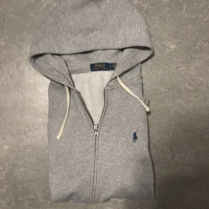 Grå hoodie från Polo Ralph Lauren - Snygg grå hoodie från Polo Ralph Lauren med dragkedja och huva. Tröjan har vit snörning i huvan och klassisk blå broderad logga på bröstet. Tillverkad i mjuk bomull, perfekt för chill dagar. Regular fit och långärmad modell.