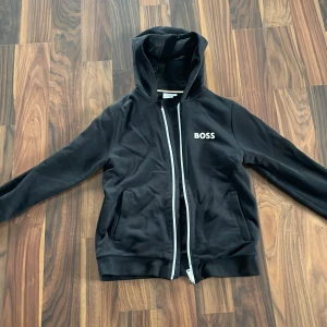 Svart hoodiejacka från BOSS XS - Svart hoodiejacka från BOSS i storlek XS. Jackan har dragkedja framtill, huva och två fickor. Snyggt BOSS-tryck på bröstet och ribbade muddar. Perfekt för en chill och stilren look.