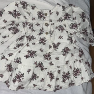 Vit blommig bebis klänning med volang - Supersöt vit kortklänning med lila blommönster, långa ärmar och volang nertill. Klänningen har knappar framtill och är sydd i mjuk manchester. Perfekt för dig som gillar romantisk och vintageinspirerad stil. Oanvända 