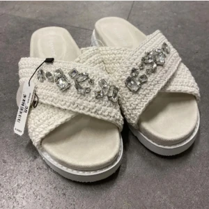 Inuikii  - Snygga vita sandaler med breda, virkade band och stora glittrande pärldetaljer. Skorna har öppen tå och platt sula, perfekta för en trendig sommarlook.