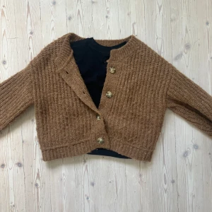 Brun stickad kofta från Zara - Mysig brun stickad kofta från Zara med grov struktur och snygga knappar framtill. Koftan har lång ärm och en croppad passform som ger en trendig look. Perfekt att slänga över en t-shirt eller topp för extra värme. Använd 1 gång.