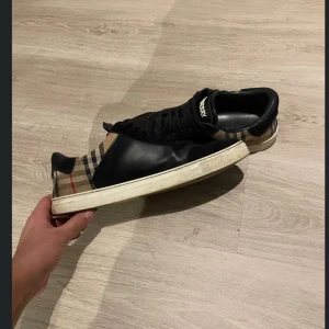 Burberry rutig sneaker i skinn - Snygga sneakers från Burberry med klassiskt rutigt mönster på sidorna och svart skinn framtill. Skorna har svarta snören, vit sula och Burberry-logga på plösen. Perfekt för dig som vill ha en lyxig och tidlös look. Kvitto finns! Köpt på farfech