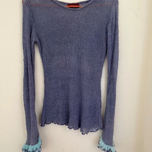 Ljuslila stickad topp från Adriana - Supersöt ljuslila stickad topp från Adriana med långärmade ärmar och volangdetaljer i ljusblått vid ärmsluten. Tunt och luftigt material i 100% rayon, perfekt för lager-på-lager. Snygg vågig kant nertill och vid ärmarna.